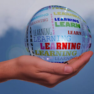 A hand is holding a clear ball against a blue sky background. The ball has "Learning" in different colors and fonts all over it.