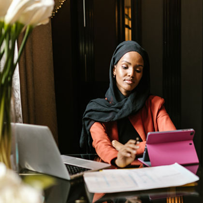 Woman of color wearing coral blazer and black head scarf looking at tablet device with a pink cover. Next to the tablet on the table are papers and an open silver laptop. There is a blurred vase of flowers in the foreground.