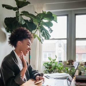 African-American woman with short curly hair wearing grey blazer and white shirt, is smiling and waving at her laptop screen. She is working at a light wood desk with windows behind her. There is a large house plant in the corner and a few small plants in the window.