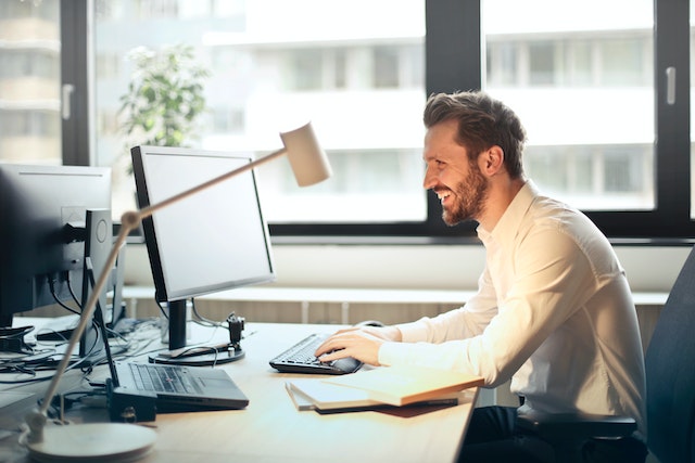 Man with beard smiles while working at a computer in an office. There is a desk lamp and a plant on the desk, and he is next to a large window.
