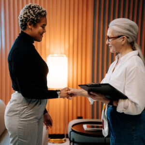 A smiling woman with long grey hair in a low ponytail and wearing glasses, a white long-sleeved button-up shirt, and jeans is holding a clipboard and shaking hands with a smiling woman of color with short dark and blonde hair wearing a long-sleeved black turtleneck and khaki pants. In the background is a paneled orange wall and a floor lamp emitting soft light.