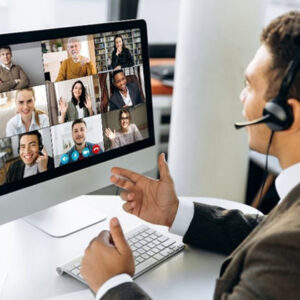 Man in business suit and wearing a headset is in front of a computer screen showing 9 people in a remote meeting.