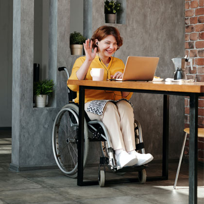A woman with short auburn hair, wearing off-white pants and a yellow-orange shirt, is sitting in a wheelchair at a table with black metal legs and a wood top. She is wearing headphones, is smiling, and waving at the laptop screen. Behind her is a grey cement wall with openings that have plants in them. The floor is made of square grey-marbled cement.