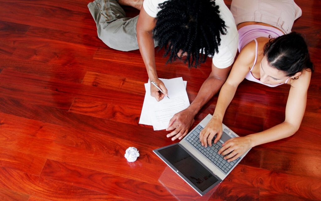 Young woman and young African-American man casually sitting on wooden floor. Woman is typing on laptop, man is writing on paper. There is a crumpled piece of paper by the laptop.