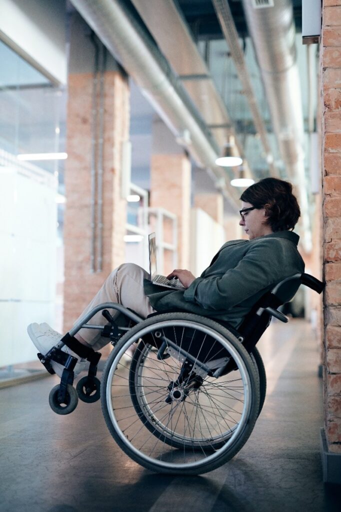 woman with short brown hair in khakis, sneakers, and jacket, wearing glasses, sitting in wheelchair leaning at an angle against a brick wall in a hallway with brick pillars, looking at open laptop on her lap