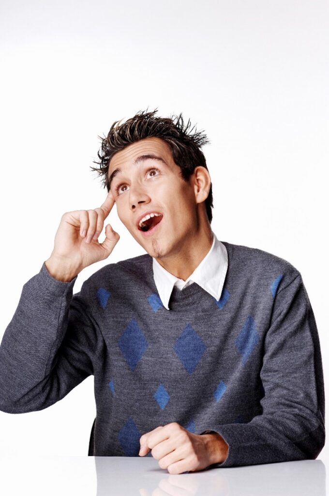 young man with short spikey hair wearing blue and grey argyle sweater pointing at forehead with “aha!” look on his face.