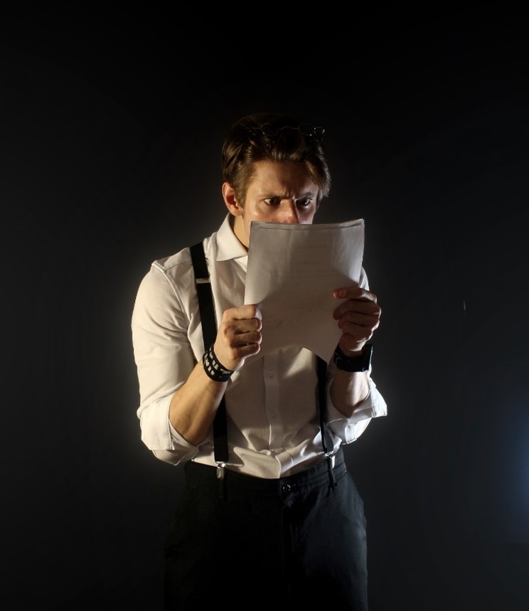 Standing young Caucasian man with brown, slightly messy hair, wearing black trousers, white long-sleeved button-up shirt, and black suspenders holding document close to his face, starting at it intently. He looks a little confused