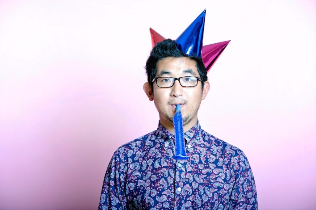 Asian man wearing black eyeglasses and three metallic party hats on his head (red, blue, and purple) in blue-purple paisley button-down shirt looking at camera. He’s blowing into a party horn and is standing in front of a vertical (top-down) white-to-light-purple gradient