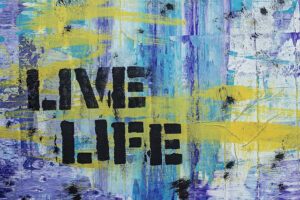 "Live Life" is written in block letters in black on a wall. The background is abstract brush strokes of yellow, light blue, purple, and white.