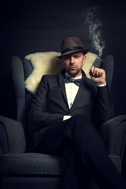 A man wearing a black suite with a black bowtie and a brown and black fedora sits in a black wingback chair. He has a serious look on his face, looking at the camera. He's smoking a cigar.