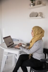 A woman wearing black pants, grey sweater and light tan headscarf is sitting at a white table typing on a laptop.