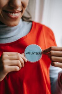Smiling woman wearing grey turtleneck under orange t-shirt putting on a large white "Volunteer" pin.
