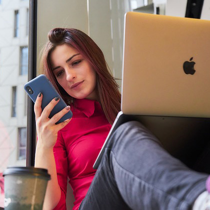 Young woman with long brown hair wearing long-sleeved button-down red shirt, jeans and sneakers sitting with MacBook on her knees while looking at cell phone. She’s sitting in front of a large window with a view of a large office or apartment building, and there is a cup of a hot beverage next to her.