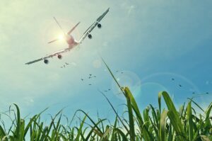 Airplane flying with the sun behind it in a bright blue sky with grass in the foreground