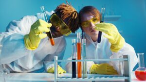 Two people wearing white lab coats, safety goggles and yellow gloves holding test tubes containing orange and dark blue liquid