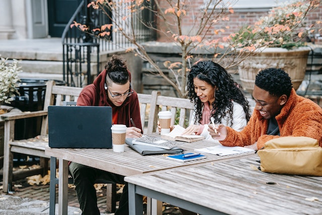 Three young cheerful ethnic people sitting at an outside table. On the table are coffee cups, a laptop, pens, and notebooks.