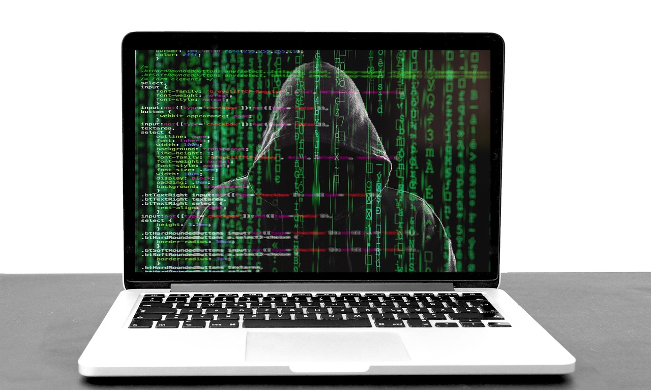 Laptop with green code on black screen with menacing figure in dark hoodie in the screen background.