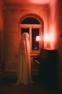 Spooky room lit in soft red with a ghost in a sheet standing at an old piano.
