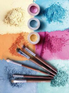 Brightly colored powders in background (orange, pink, blue, aqua, yellow) with eyeshadow pots and brushes in foreground
