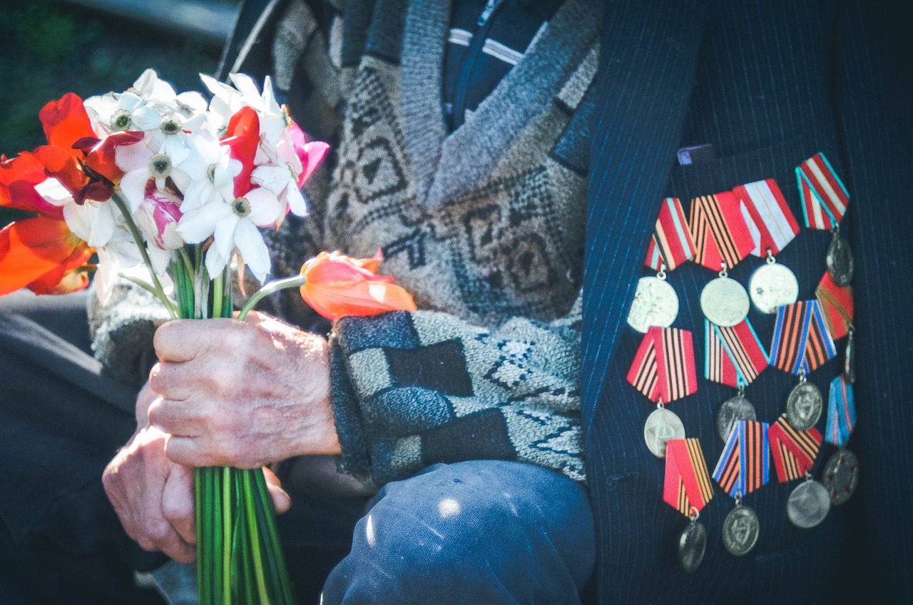 Older veteran with several medals on jacket, holding flowers.