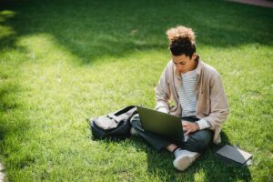 Young person wearing jeans and sweater sitting on the grass, working on a laptop.