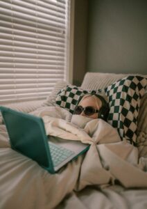 Person under covers wearing a headset and dark sunglasses, working on computer