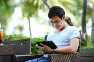 Young man outside at table looking at tablet and smiling.