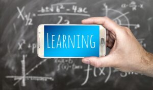 Hand holding phone with "LEARNING" on the screen in front of slightly blurry math equations on blackboard.