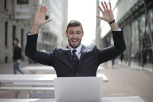 Young man in suit happily raising hands while working on a laptop at an outside table in the city.