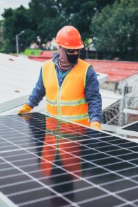 Person wearing orange safety vest and hardhat, black face mask installing solar panel.