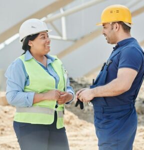 Woman wearing white hardhat and yellow safety vest talking to man wearing yellow hardhat in construction zone.