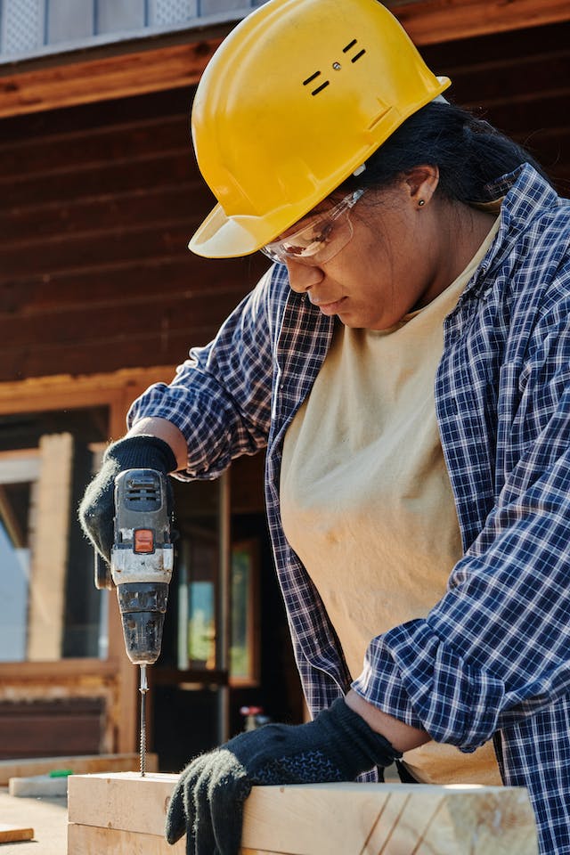 Woman wearing yellow hardhat, safety glasses, tan t-shirt, and blue plaid over shirt using power drill.
