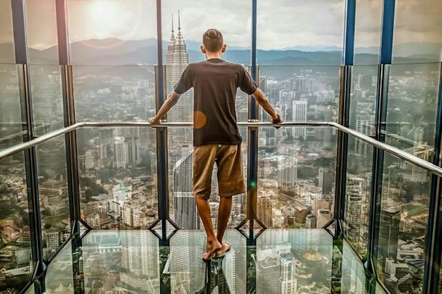 Man wearing shorts and t-shirt in glass elevator looking forward over a cityscape and mountains.