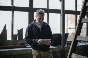 Grey-haired bearded man in black hoodie sweatshirt and khaki pants standing in front of large window in an old building using tablet device.