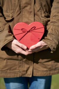 Person wearing dark khaki jacket holding a red heart-shaped box with a small bow on it.