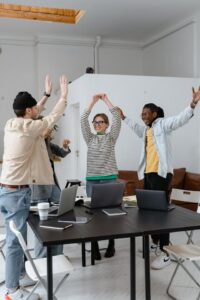 Group of four people standing around a table with laptops on it. They are celebrating with smiles and hands in the air.