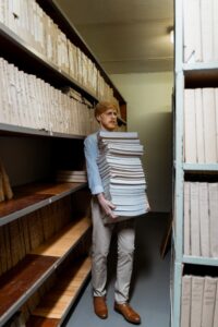 Confused-looking man carrying a large stack of documents into a room full or more documents on shelves.