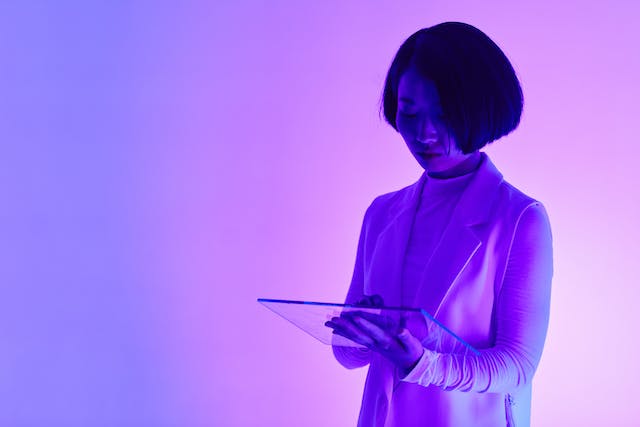 Woman with short dark hair and wearing a white suit is using a clear tablet. The background of the picture is blue to purple gradient.