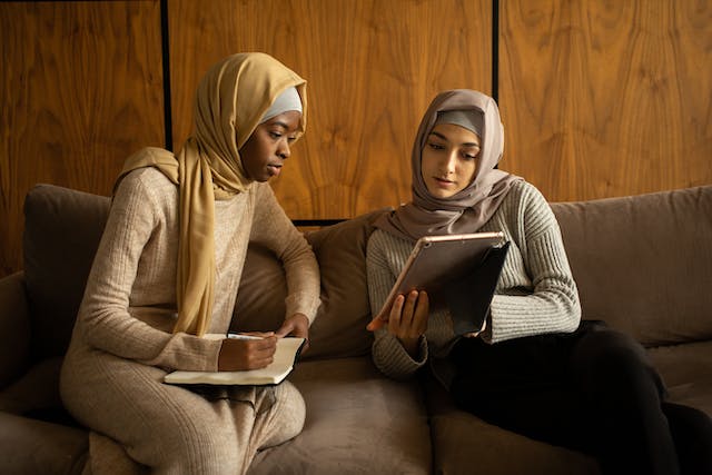 Two women wearing light clothes and head coverings sit on couch looking at a tablet.