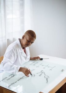 Woman wearing white lab coat writing equations on a large clear board.