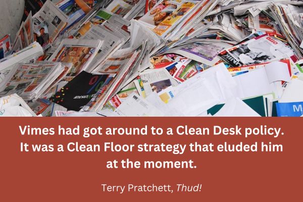 Pile of papers and magazines with text: “Vimes had got around to a Clean Desk policy. It was a Clean Floor strategy that eluded him at the moment. – from Terry Pratchett’s Thud!” 