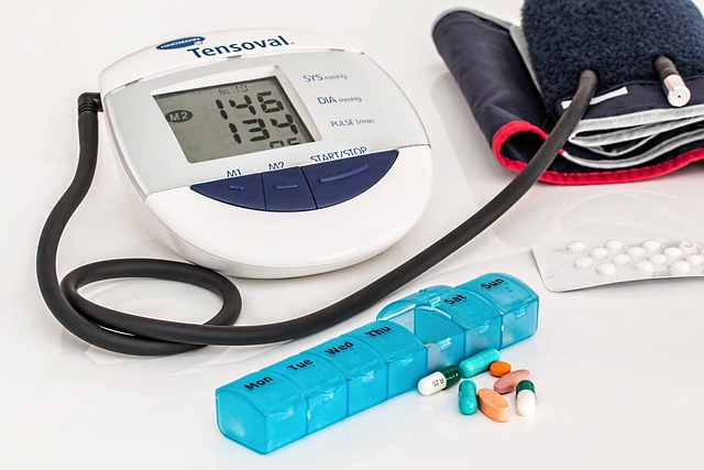 There is a blood pressure cuff, a silver pack of white pills, a blue pill box with daily compartments, and some pills lying on a white surface.