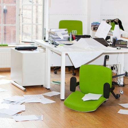 Office with white walls, desk, and cabinets, and a light wood floor. A lime green office chair is on both sides of the desk; one is on its back. Papers are strewn around the floor and desk.
