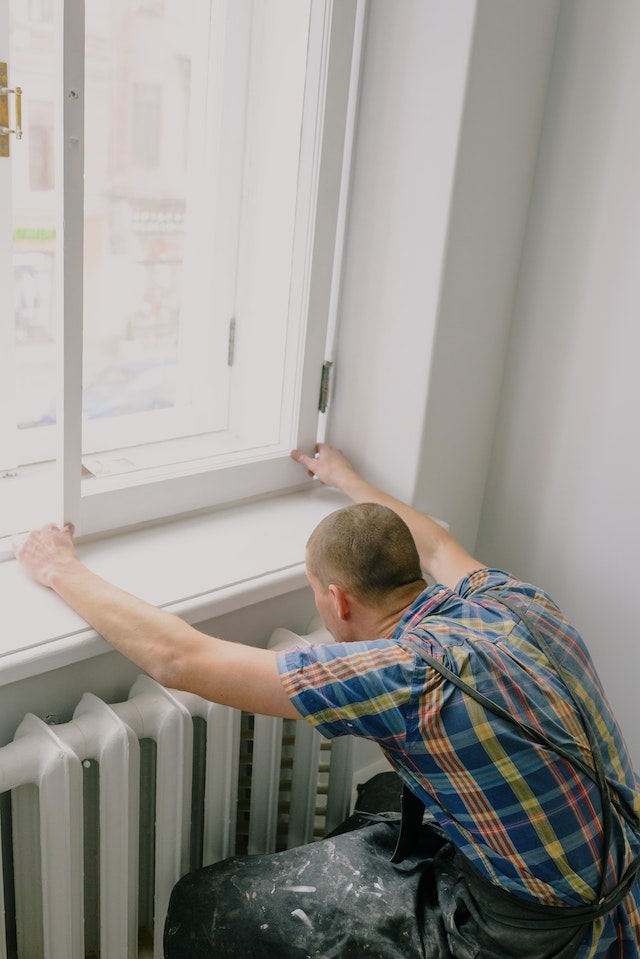 A man in a plaid shirt and a work apron is bending down to carefully ensure he’s properly installing a window.