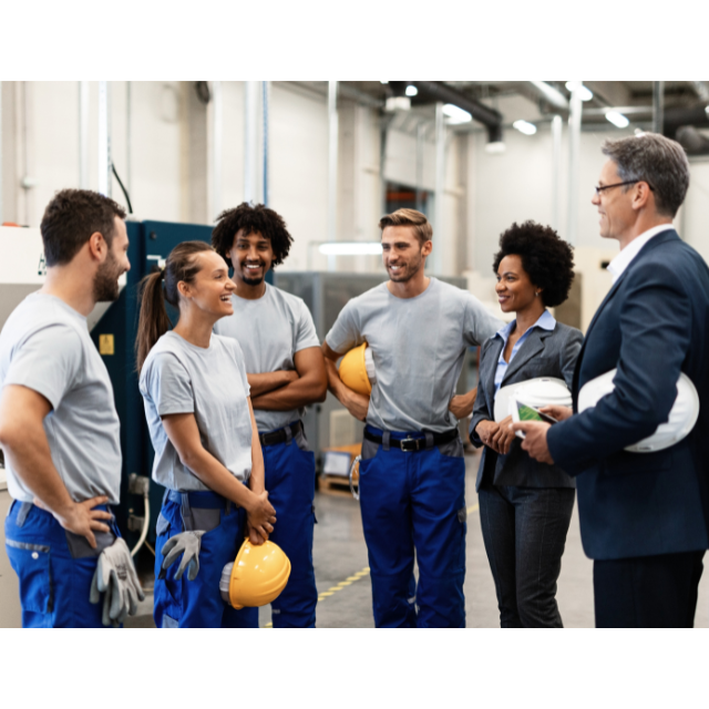 Four smiling people in matching blue and grey uniforms and yellow hardhats talking to two smiling people wearing suits with white hardhats.
