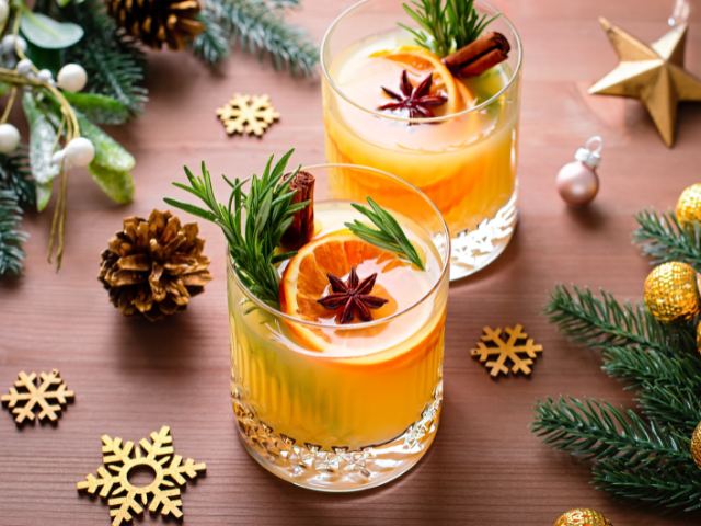 Cidery drinks topped with orange slices and cinnamon sticks sitting on a table that has greenery and bold snowflake, star, and round ball ornaments scattered on it.