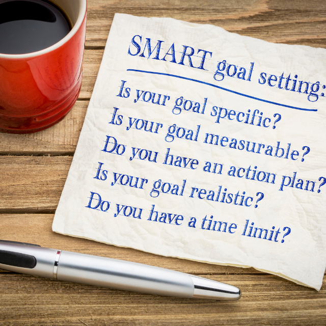Coffee mug and pen on a wooden table, along with a note about SMART goal setting. Caption reads: "The reason that goals are more beneficial than resolutions is that goals are more specific and action oriented than resolutions, which makes it easier to create a plan to achieve them." - Ashira Prossack, internationally recognized communication coach and trainer