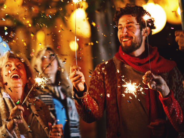 Smiling people in warm clothes standing outside holding sparklers while sparkly confetti is in the air.