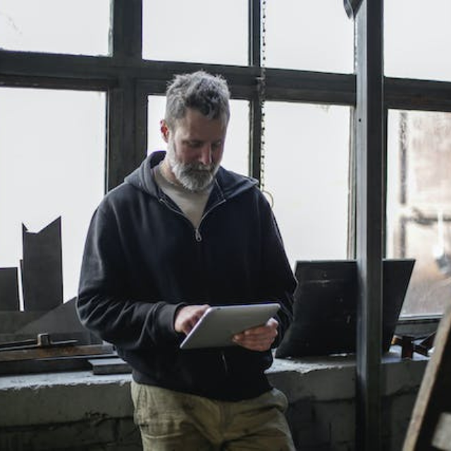 A man with gray hair and a beard stands indoors by large industrial-style windows, looking down at and using a tablet. The setting appears to be a workshop or studio with natural light streaming in.