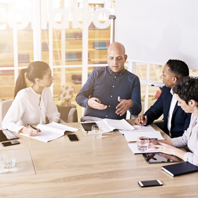 Four professionals sit around a conference table reviewing documents while one person leads the discussion, gesturing as they speak during a team meeting in a modern office.